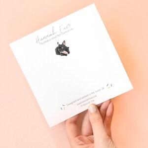 May include: A square white greeting card featuring a small illustration of a black French Bulldog holding a pink and white striped treat. The text reads Hannah Farr Illustrator and Pet Portraiture at the top and Designed and printed in the lovely UK at the bottom with the website and copyright information.