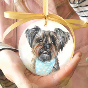 Op de afbeelding: Een handgeschilderd keramiek ornament met een portret van een kleine, bruin-zwarte hond die een lichtblauwe bandana met witte stippen draagt. Het ornament is vastgebonden met een gouden lint.