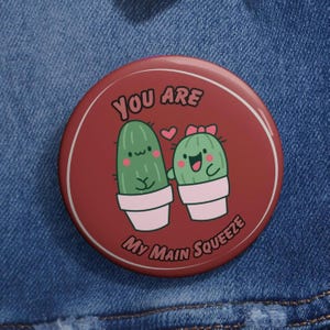 May include: A round, maroon button with the text "YOU ARE My Main Squeeze" encircling two cartoon cacti in white pots. One cactus has a bow, and both have smiling faces. The background is a denim fabric.