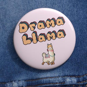Può includere: Un bottone rotondo rosa chiaro con le parole "Drama Llama" in lettere arancioni, turchesi e rosse. Sotto il testo c'è un lama dei cartoni animati con una coperta colorata e una sciarpa rosa. Il bottone è su uno sfondo di jeans blu.