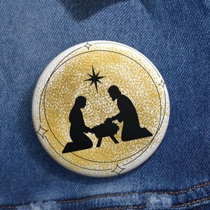 May include: A round, white button with a gold glitter background and a black silhouette of the Nativity scene. The button has a black outline and star accents.