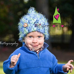 May include: A child wearing a blue and green knitted hat with a wooden button. The child is also wearing a blue sweater. The background is blurred, with the text "Light Bender Photography" and "whisper from your Heart".