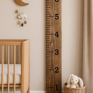 May include: A wooden height chart with black markings and numbers, measuring up to 183 cm. A wicker basket holds a stuffed bunny. A wooden crescent moon and cloud mobile hangs above a wooden cot.