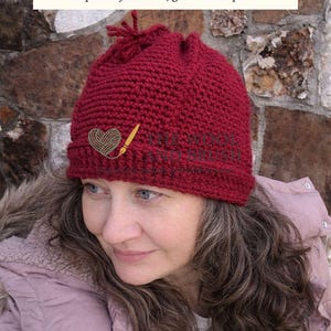 May include: A deep red, handmade crochet hat with a drawstring closure. The hat features a small embroidered heart with yarn and a paintbrush design. The text "THE WOOL AND BRUSH" is visible above the hat, with the tagline "Inspired by creation, grounded in peace."