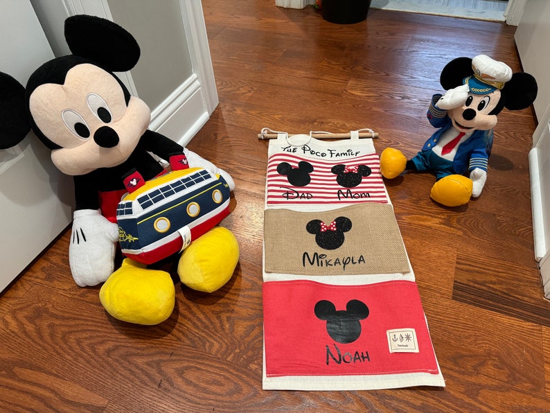 Pu&ograve; includere: Un organizzatore da parete in tela bianca con quattro tasche che presentano silhouette di Topolino e il testo "The Poco Family", "Dad", "Mom", "Mikayla" e "Noah".