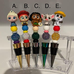 May include: Five colorful bottle stoppers with different cartoon characters on top. The characters are Elsa from Frozen, Jessie from Toy Story, Maui from Moana, a white puppy, and Olaf from Frozen. The stoppers are on a clear plastic stand.