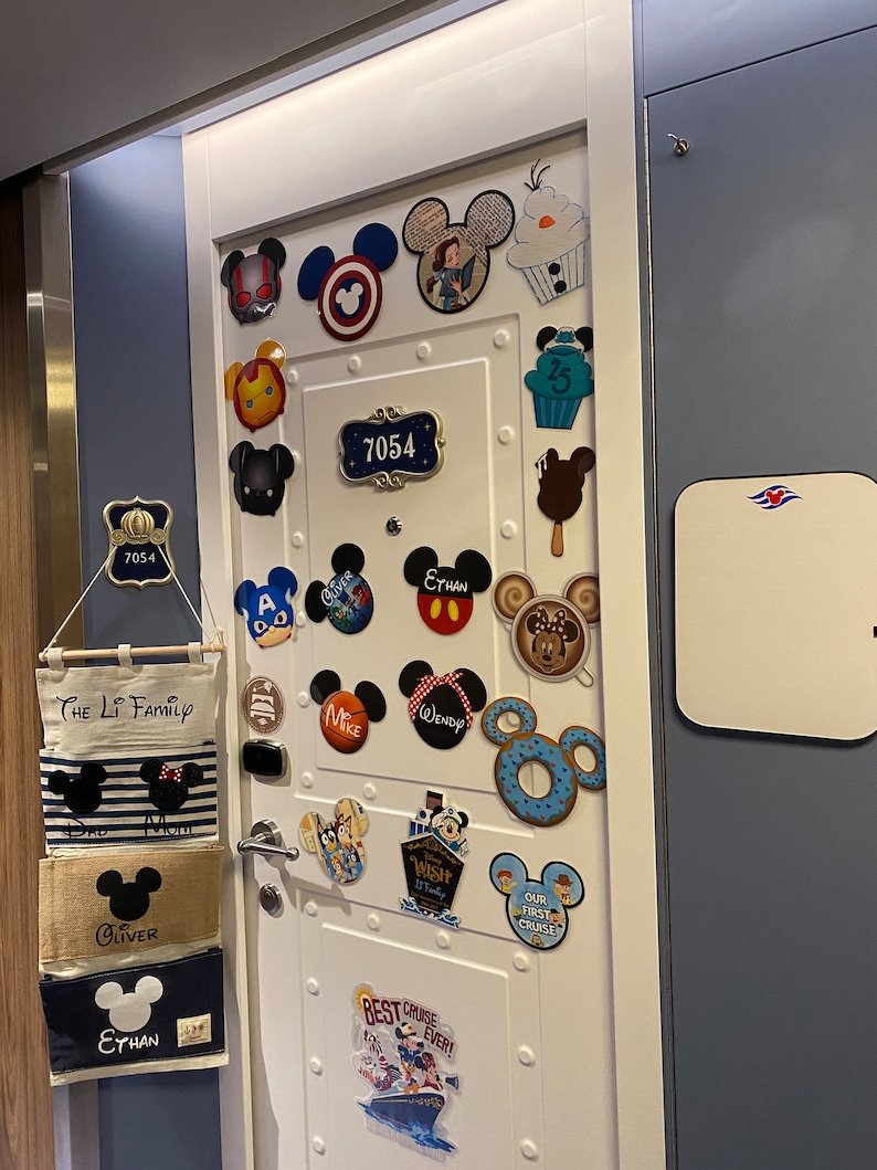 Pu&ograve; includere: Una porta bianca con una variet&agrave; di magneti e adesivi a tema Disney. La porta ha una piccola lavagna bianca con un logo blu e bianco. La porta ha anche una targa con il numero "7054".