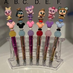 May include: Six colorful pens with different cartoon character toppers. The pens are displayed on a clear plastic stand. The toppers include a yellow chick, a brown dog, a purple cat, a brown bird, a pink cat, and a brown bear.