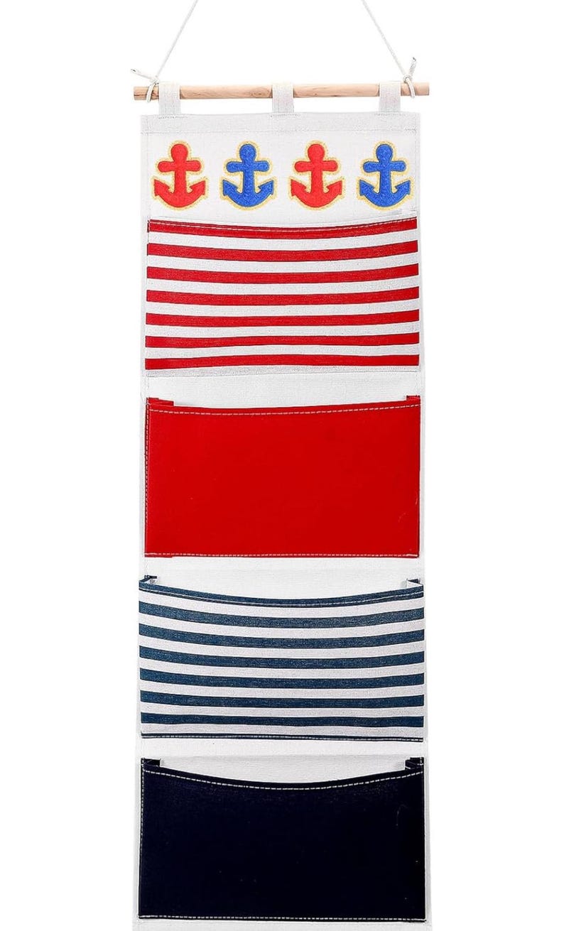 Pu&ograve; includere: Un organizzatore da parete in tela bianca con quattro disegni di ancore blu e rosse. L'organizzatore ha tre tasche con strisce rosse e bianche, strisce blu e bianche e tessuto rosso tinta unita.