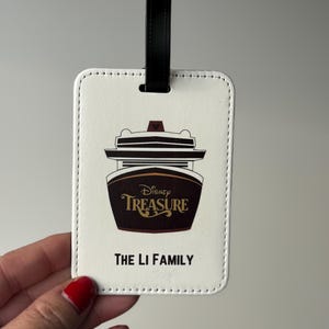 May include: White luggage tag with a black leather strap. The tag features a cartoon image of the Disney Treasure cruise ship with the text "Disney Treasure" and "The Li Family".