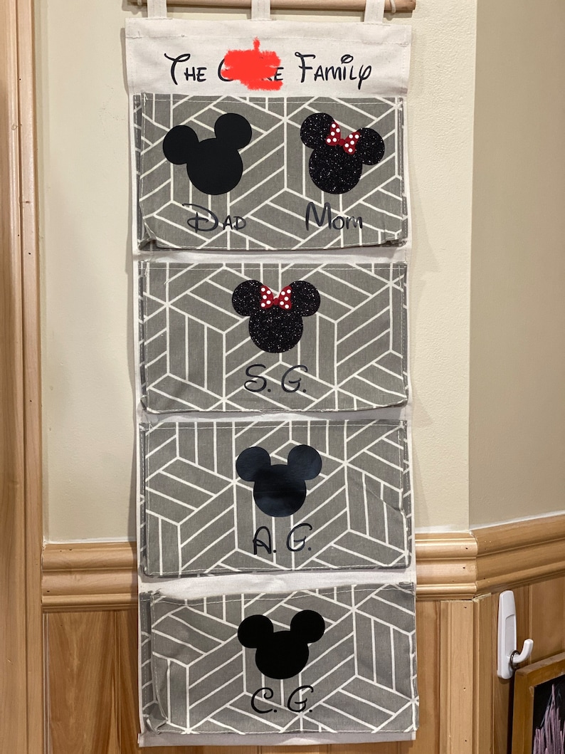 Pu&ograve; includere: Un organizzatore da parete in tela bianca con quattro tasche. Ogni tasca presenta un motivo geometrico grigio e una silhouette di Topolino nera. La tasca superiore ha il testo "The C Family" scritto in nero. Le tasche sono etichettate "Dad", "Mom", "S.G." e "C.G."