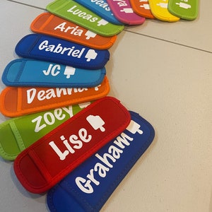 May include: A set of 12 colorful neoprene can coolers with personalized names, including Jesus, Lily, Sonia, Arria, Lucas, Gabriel, JC, Deanna, Zoey, Lise, and Graham. Each cooler features a white thumbs up icon.
