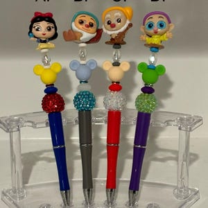 May include: Four pens with different colored rhinestones and Disney character toppers. The toppers are Snow White, a dwarf, a dwarf, and a purple character. The pens are displayed in a clear plastic stand. The letters A, B, C, and D are above each pen.