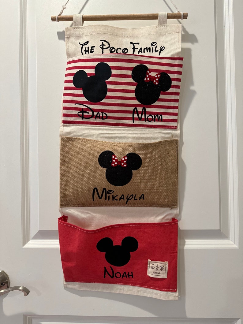 Pu&ograve; includere: Un organizzatore da parete in tela bianca con quattro tasche. La tasca superiore ha il testo "The Poco Family" e presenta silhouette di Topolino e Minnie Mouse. La seconda tasca ha il testo "Dad" e "Mom". La terza tasca ha il testo "Mikayla" e la quarta tasca ha il testo "Noah".