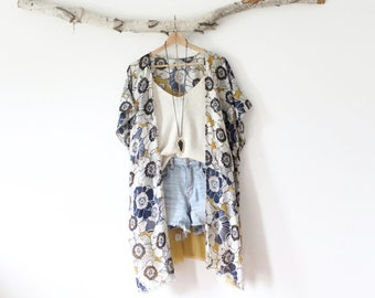 May include: A floral kimono with navy, white, and mustard yellow patterns layered over a cream-colored tank top and light blue denim shorts. A long necklace with a black arrowhead pendant hangs over the tank top, all on a wooden hanger.