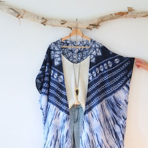 May include: A blue and white tie-dye kimono with fringe trim. The kimono is hanging on a wooden branch and is layered over a white top with a gold pendant necklace.