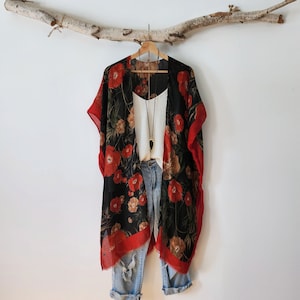 May include: A black kimono featuring a red floral print with a wide red border, layered over a white sleeveless top and light blue distressed denim jeans. A long necklace with a pointed pendant hangs over the white top.