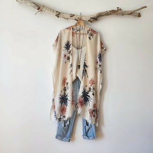 May include: A cream-colored kimono with a floral print in shades of blue and rust, layered over a white tank top and light-wash distressed denim jeans. A long necklace with a black arrowhead-shaped pendant hangs over the tank top.
