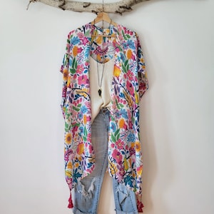 May include: A white kimono with a floral print in shades of pink, blue, yellow, and green. The kimono has tassels on the bottom and is hanging on a branch. It is paired with blue jeans.