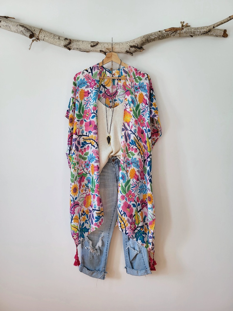 May include: A white kimono with a floral print in shades of pink, blue, yellow, and green. The kimono has tassels on the bottom and is hanging on a branch. It is paired with blue jeans.