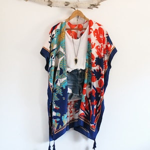 May include: A kimono-style cover-up featuring a vibrant floral pattern in shades of navy blue, bright orange, and teal, finished with navy blue tassels at the hem. It is layered over a plain white tank top, denim shorts, and a long arrowhead pendant.