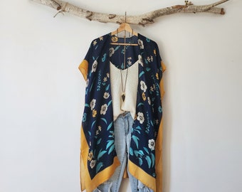 May include: A navy blue kimono with a floral pattern in white, yellow, and teal, featuring a mustard yellow border and tassels. It is layered over a white camisole and light-wash distressed denim jeans, with a long necklace featuring a dark pendant.