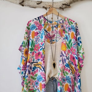 May include: A white kimono with a colorful floral print. The kimono has short sleeves and a long, flowing silhouette. It is hanging on a wooden branch.