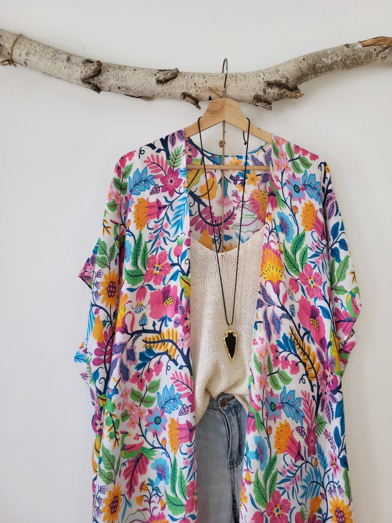 May include: A white kimono with a colorful floral print. The kimono has short sleeves and a long, flowing silhouette. It is hanging on a wooden branch.