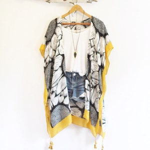 May include: A black and white sunflower print kimono with mustard yellow trim and tassels, layered over a white tank top and blue denim shorts, featuring a long necklace with a black arrowhead pendant on a gold chain.