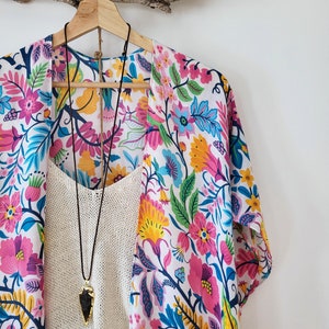 May include: A white kimono with a colorful floral print. The kimono has a long sleeve and is hanging on a wooden hanger. A cream-colored tank top and a necklace with a dark gray arrowhead pendant are visible underneath the kimono.