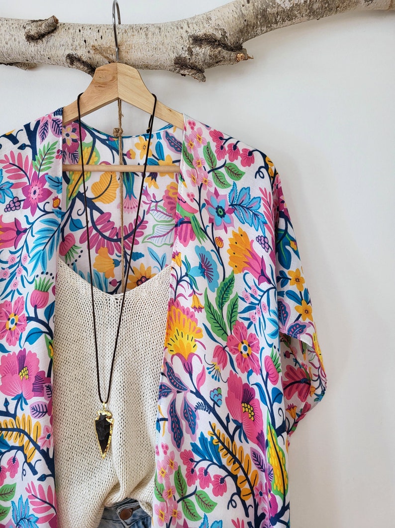 May include: A white kimono with a colorful floral print. The kimono has a long sleeve and is hanging on a wooden hanger. A cream-colored tank top and a necklace with a dark gray arrowhead pendant are visible underneath the kimono.
