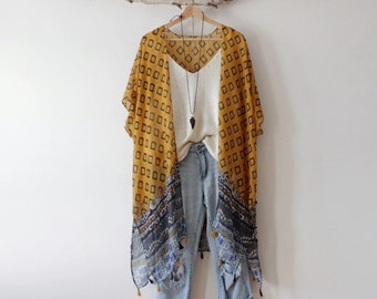 May include: A mustard yellow kimono with a black square pattern and a blue and white floral border featuring small tassels, worn over a white v-neck camisole and light wash distressed denim jeans, accessorized with a black arrowhead necklace.