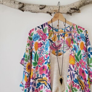 May include: A white kimono with a colorful floral print. The kimono has a wide, flowing silhouette and is perfect for layering over a simple top or dress. The kimono is adorned with a black necklace with a large, dark stone pendant.