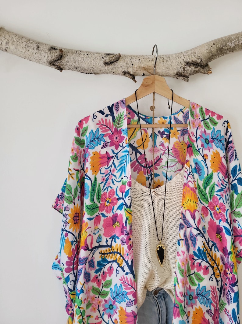 May include: A white kimono with a colorful floral print. The kimono has a wide, flowing silhouette and is perfect for layering over a simple top or dress. The kimono is adorned with a black necklace with a large, dark stone pendant.