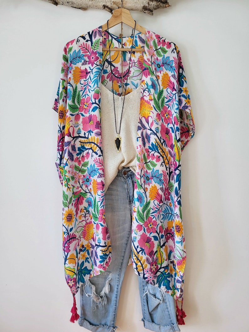 May include: A white kimono with a colorful floral print and pink tassels. The kimono is worn over a white tank top and blue jeans.