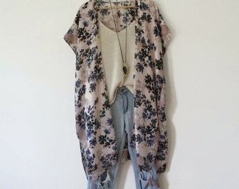 May include: A floral print kimono in shades of pink, blue, and black, paired with a cream-colored tank top and light blue distressed denim shorts. A long necklace with a pendant hangs over the tank top. The outfit is displayed on a wooden hanger.