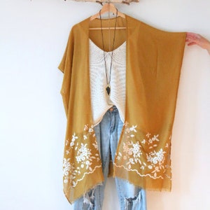 May include: A mustard yellow kimono with white floral embroidery along the hem. The kimono is hanging on a wooden branch and is styled with a white knit top and blue jeans.