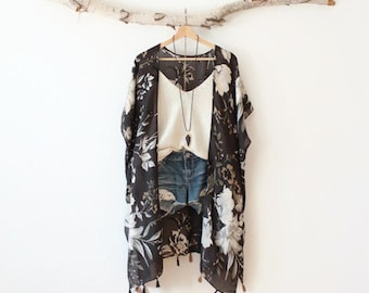 May include: A black kimono with a white floral print and small tassels along the hem, layered over a cream-coloured tank top and blue denim shorts. A long necklace with a dark pendant hangs over the tank top, all displayed on a wooden hanger.