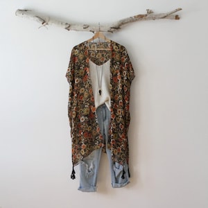 May include: A sheer kimono featuring a dark floral print with orange, yellow, and white flowers, finished with black tassels. It is layered over a white camisole and light-wash distressed denim jeans, accessorized with a long black pendant necklace.