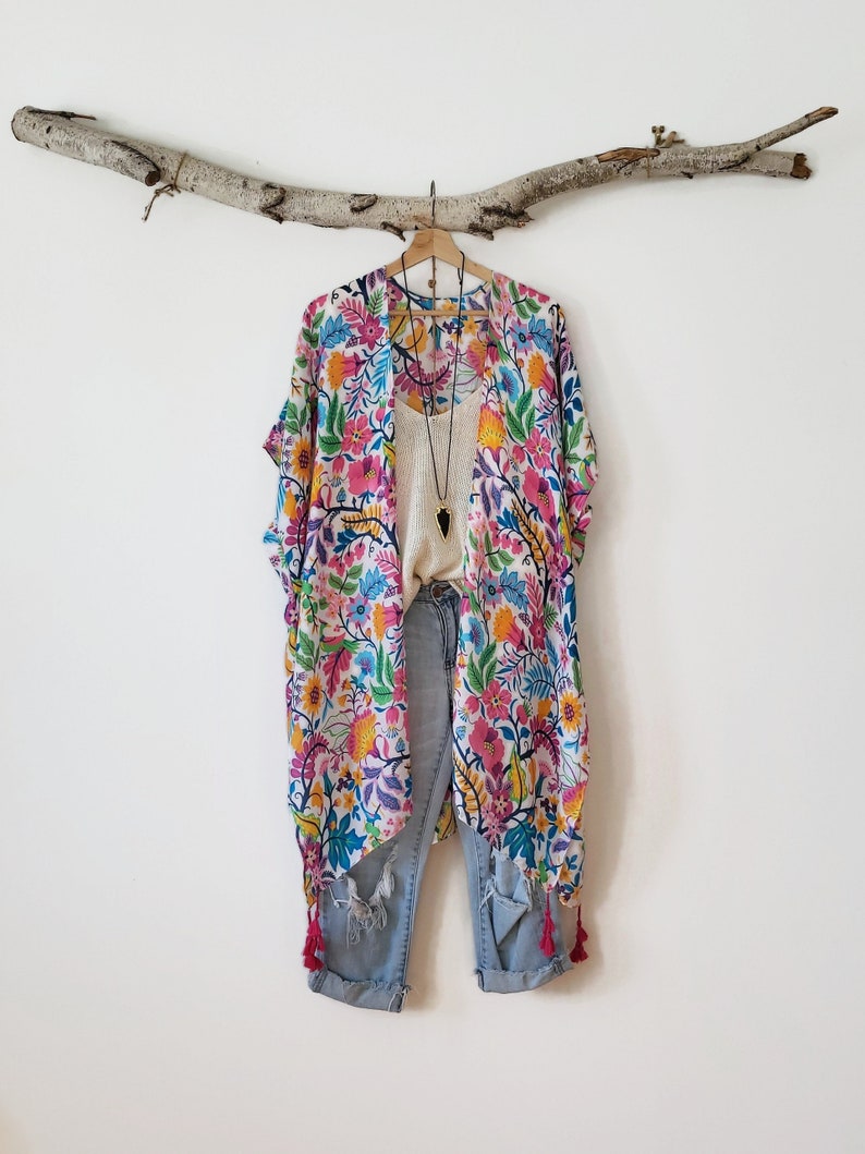 May include: A white kimono with a floral print in shades of pink, blue, yellow, and green. The kimono has tassels on the bottom and is hanging on a wooden branch.
