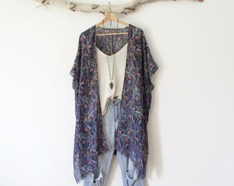 May include: A sheer, dark blue kimono featuring a multi-colored botanical leaf print, layered over a cream-colored tank top and light-wash distressed denim jeans. A long necklace with a dark, pointed pendant hangs over the tank top.