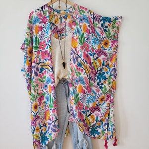 May include: A white kimono with a colorful floral print, featuring pink, blue, yellow, and green flowers. The kimono has long sleeves and a tassel trim. It is paired with blue jeans.