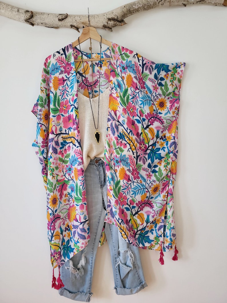 May include: A white kimono with a colorful floral print, featuring pink, blue, yellow, and green flowers. The kimono has long sleeves and a tassel trim. It is paired with blue jeans.
