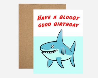May include: A birthday card with a cartoon shark illustration. The shark is light blue with a white belly and a toothy grin. The card has the text "Have a bloody good birthday" in red. Includes a brown envelope.