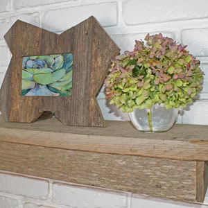 May include: A rustic wooden frame with a watercolour succulent painting, set on a weathered wooden shelf. A clear glass vase holds a bouquet of green and pink flowers. The background is a white brick wall.