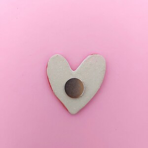May include: A white heart-shaped ceramic magnet with a silver round magnet in the center. The magnet is on a pink background.