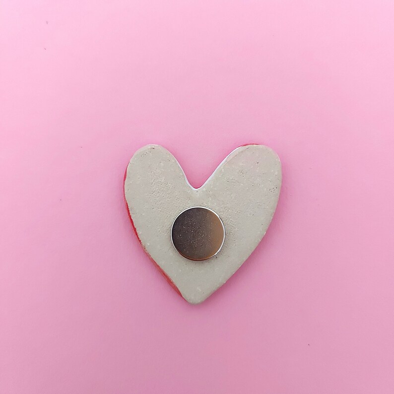 May include: A white heart-shaped ceramic magnet with a silver round magnet in the center. The magnet is on a pink background.