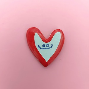 May include: A red heart-shaped ceramic ornament with a white painted interior and a blue smiley face.