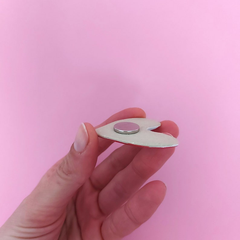 May include: A white heart-shaped magnet with a red back. The magnet is held in a hand.