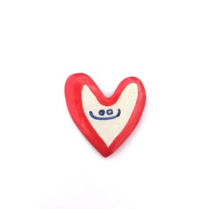 May include: A red heart-shaped ceramic ornament with a white center and a hand-painted blue smiley face.